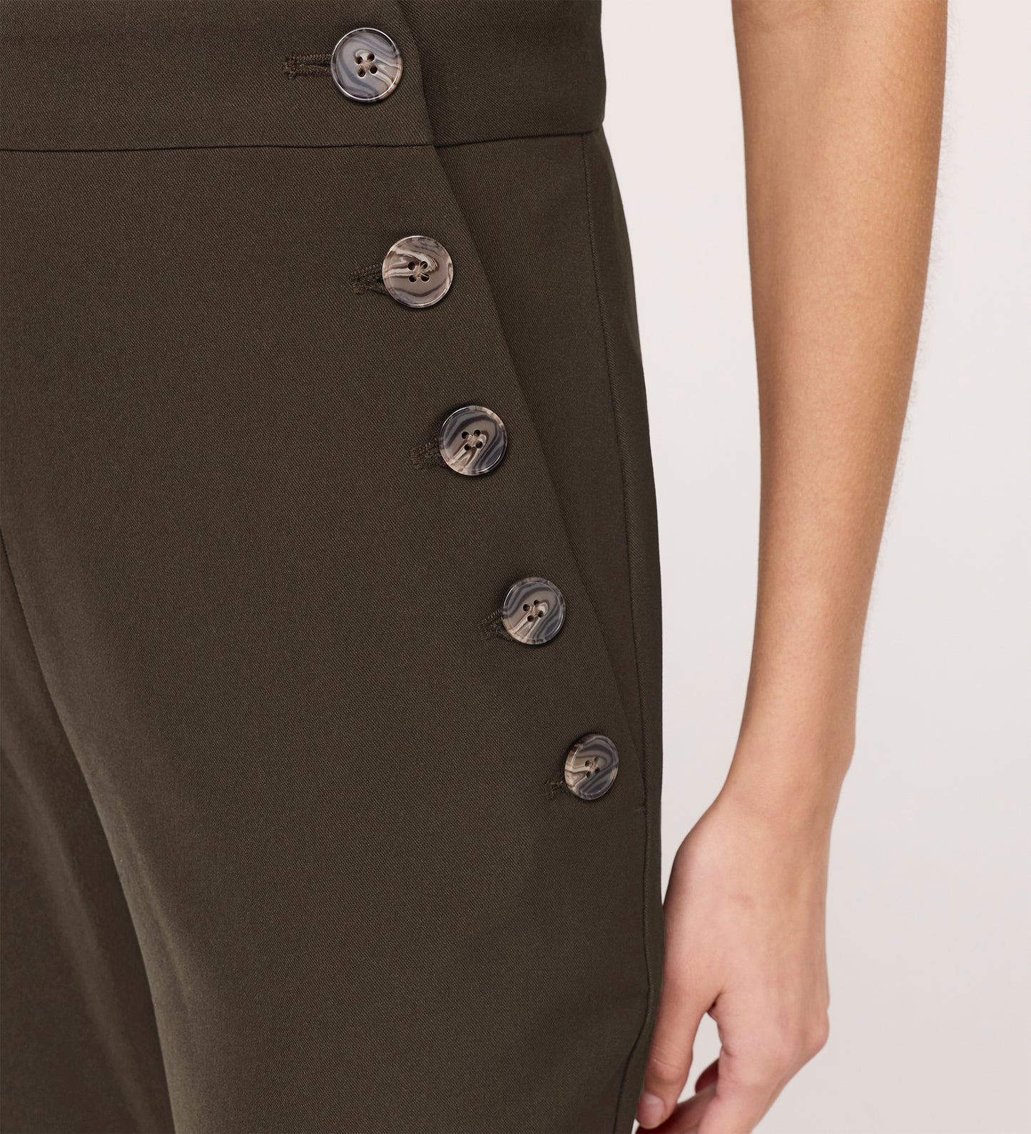 A close-up of a person wearing Nellie Mushroom Gaberdine Twill Wide Leg Trousers, high-waisted with a dark olive green hue and side row of black buttons. Their right arm and hand rest at their side.