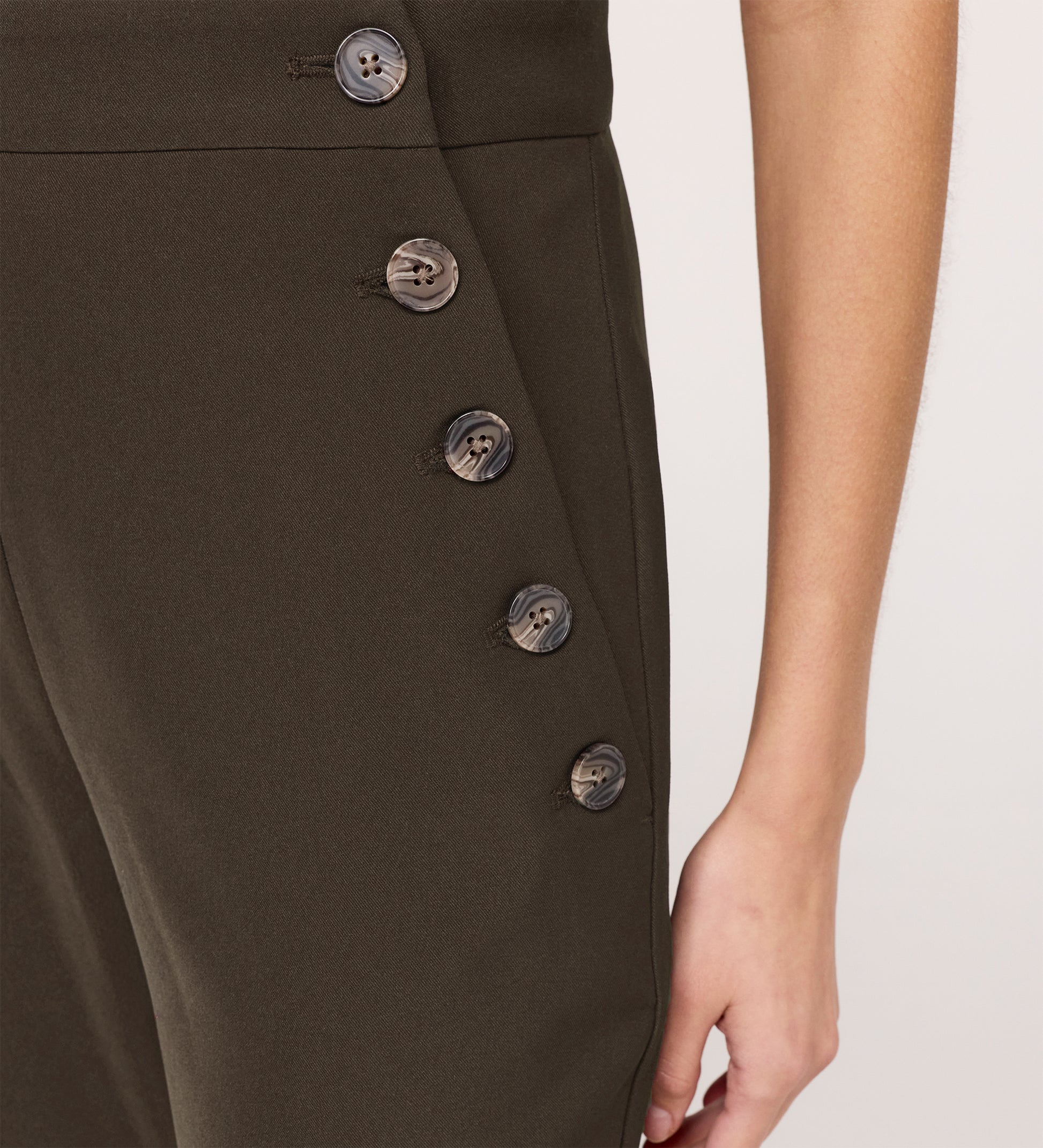 A close-up of a person wearing Nellie Mushroom Gaberdine Twill Wide Leg Trousers, high-waisted with a dark olive green hue and side row of black buttons. Their right arm and hand rest at their side.