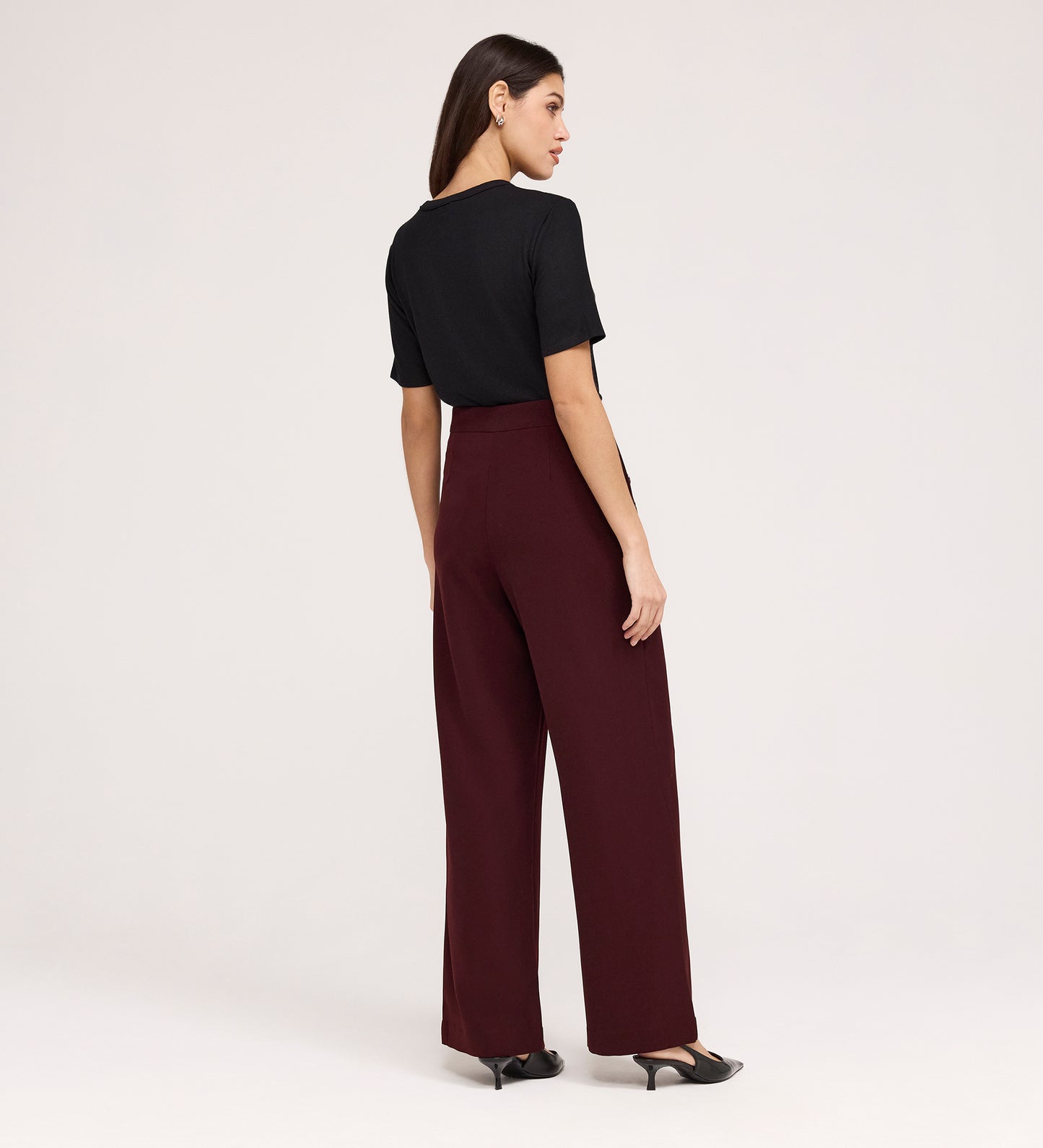 A woman, seen from behind, wears a black short-sleeve top, black heels, and Nellie Burgundy Gaberdine Twill Wide Leg Trousers against a plain light background.
