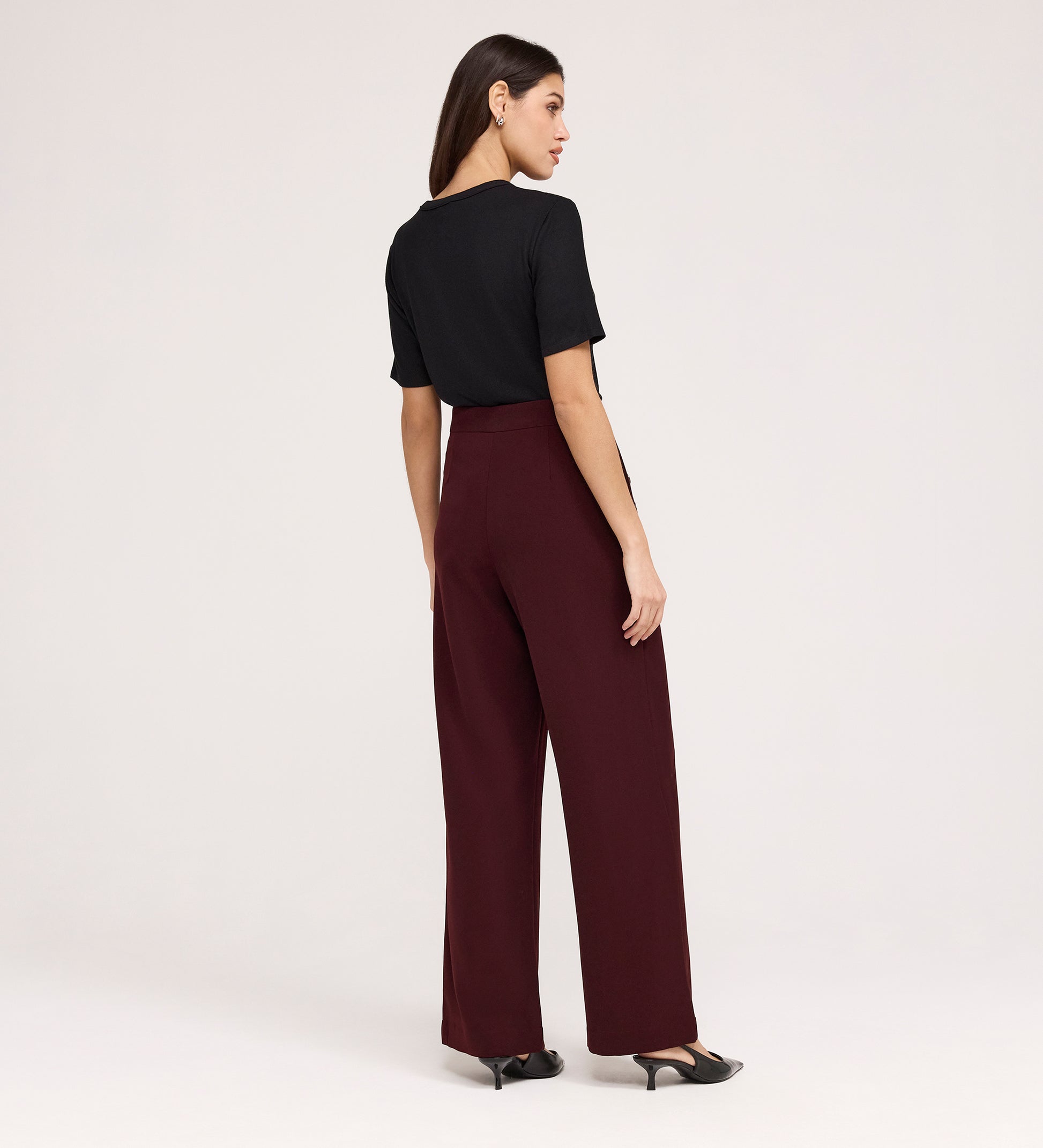A woman, seen from behind, wears a black short-sleeve top, black heels, and Nellie Burgundy Gaberdine Twill Wide Leg Trousers against a plain light background.