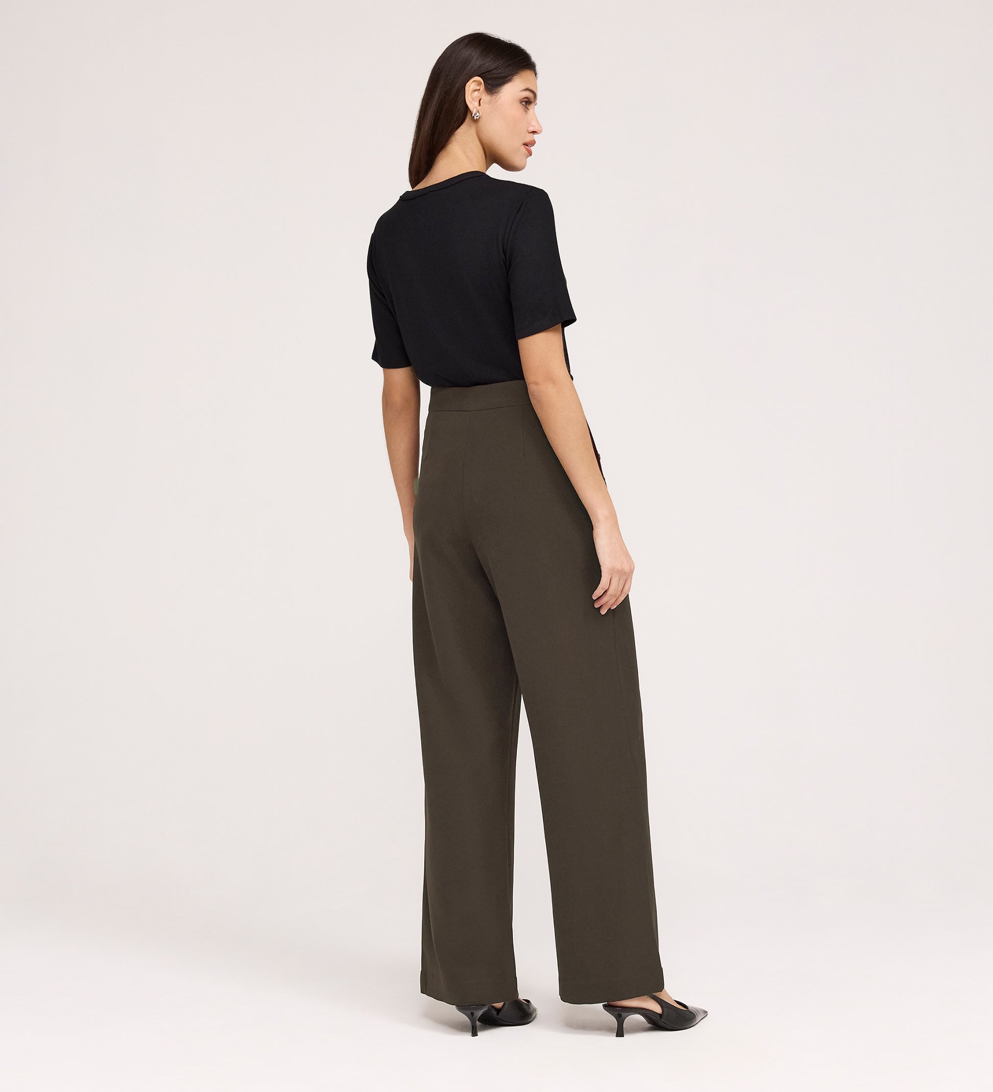 A woman stands facing away, wearing a black short-sleeve top, black low-heeled shoes, and Nellie Mushroom Gaberdine Twill Wide Leg Trousers against a plain light background.