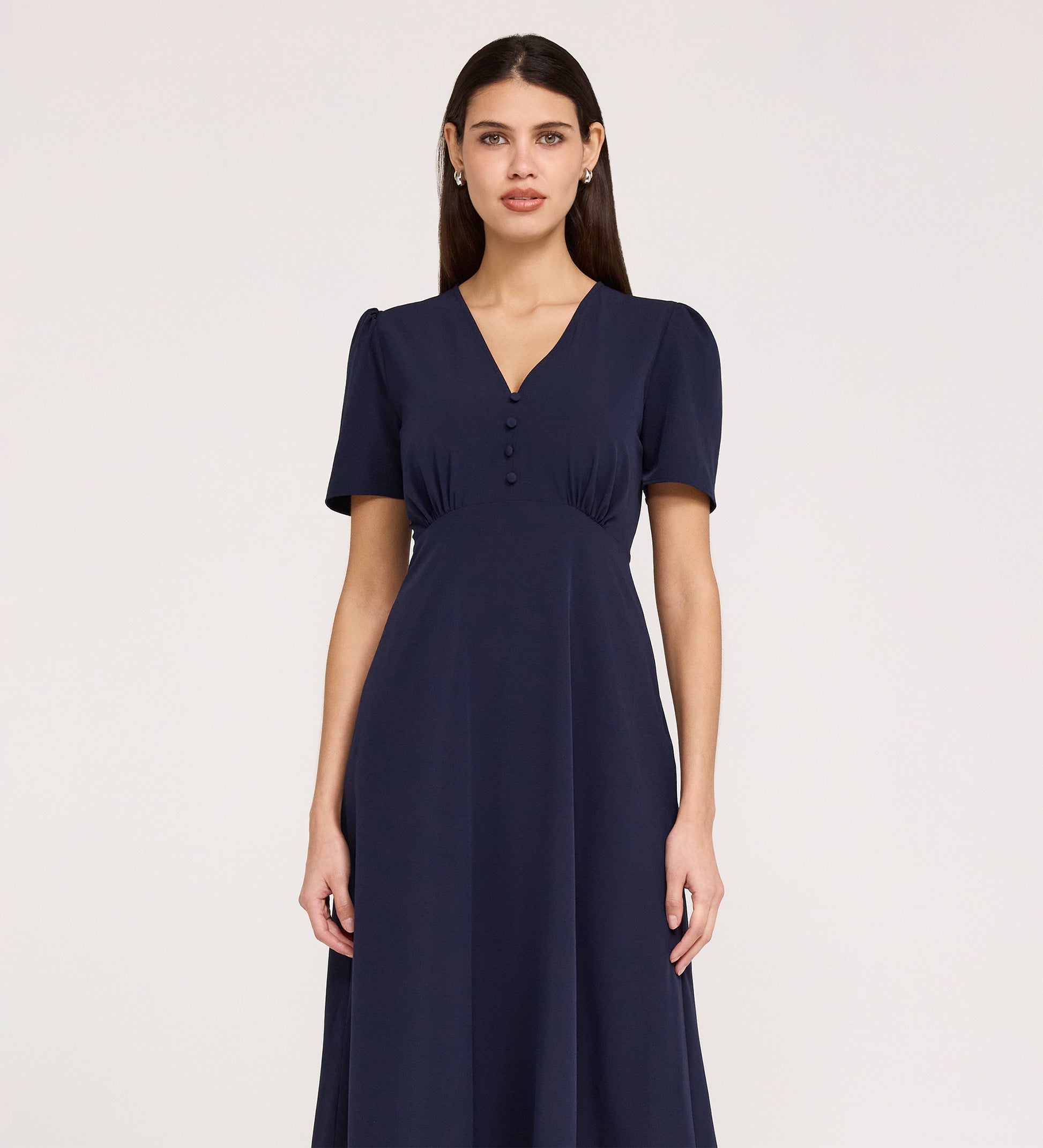 A woman wears the Nieve Navy Crepe Midi Dress, featuring short sleeves, a V-neckline, empire waist, and bodice buttons. She stands against a plain light background, facing forward with a neutral expression.