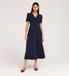 A woman stands against a plain background wearing the Nieve Navy Crepe Midi Dress, a short-sleeved, V-neck midi dress with buttons on the bodice, an empire waist silhouette, and pointed burgundy heels.
