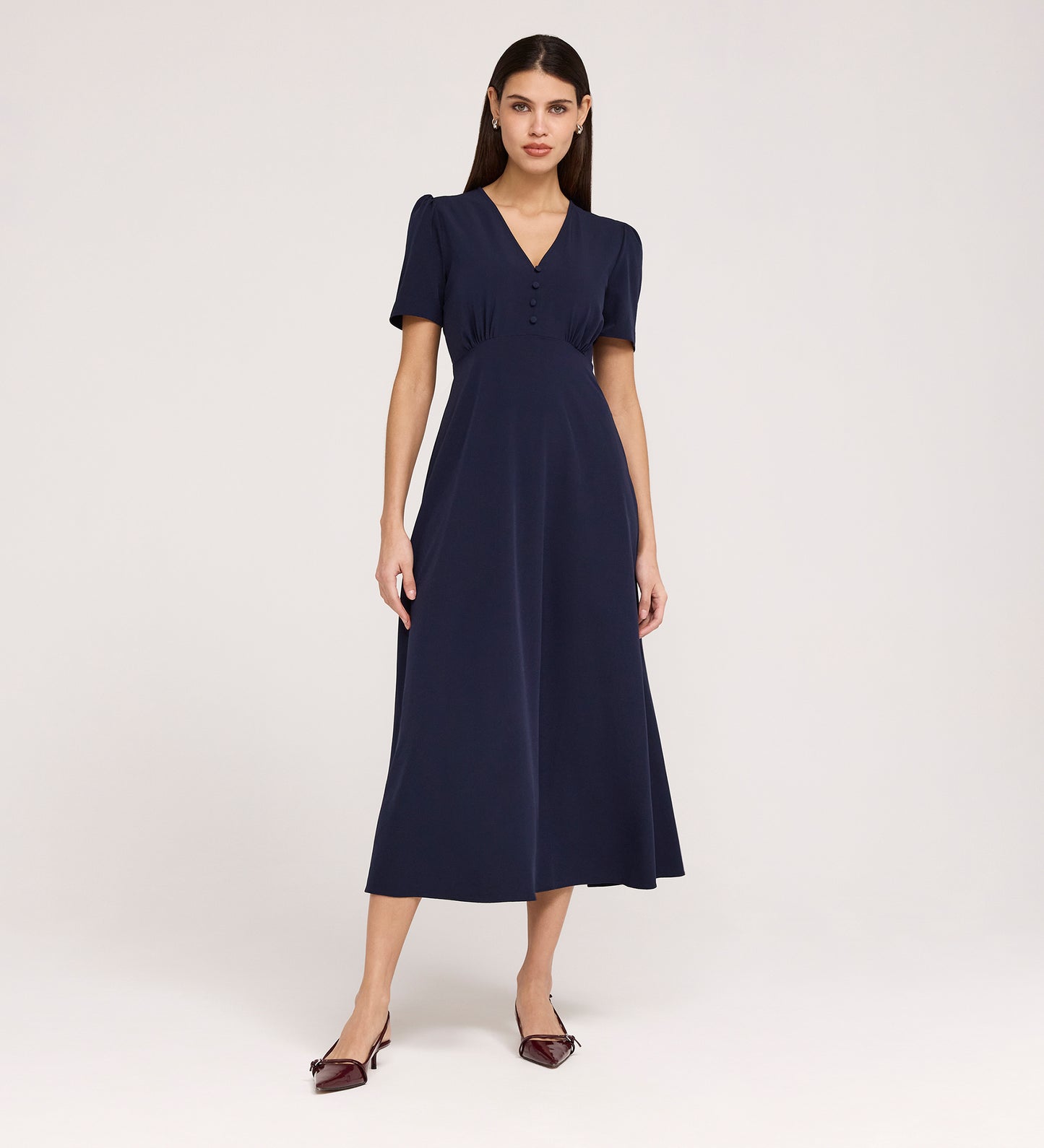 A woman stands against a plain background wearing the Nieve Navy Crepe Midi Dress, a short-sleeved, V-neck midi dress with buttons on the bodice, an empire waist silhouette, and pointed burgundy heels.