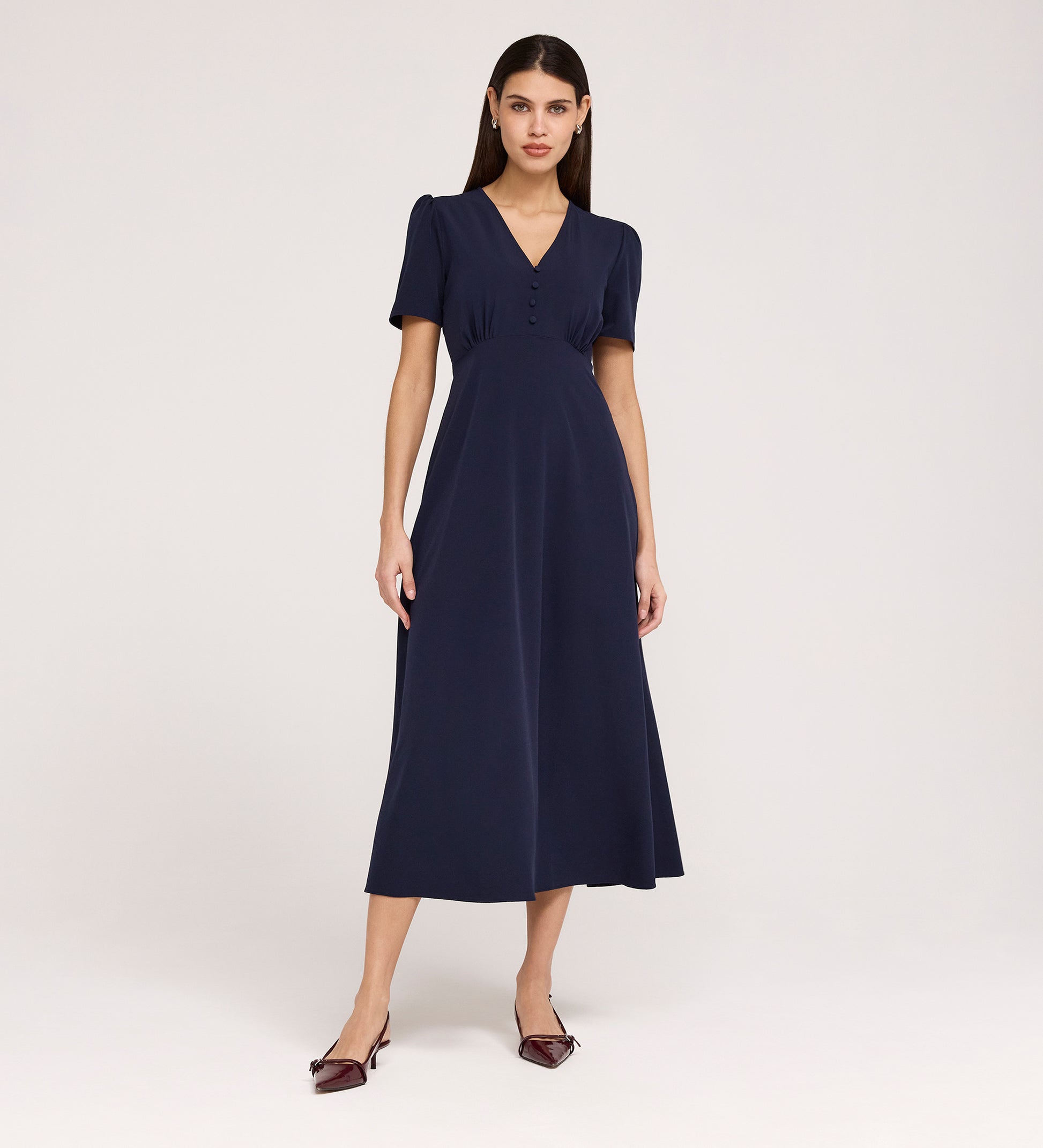 A woman stands against a plain background wearing the Nieve Navy Crepe Midi Dress, a short-sleeved, V-neck midi dress with buttons on the bodice, an empire waist silhouette, and pointed burgundy heels.