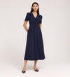 A woman with long brown hair stands against a plain background wearing the Nieve Navy Crepe Midi Dress, a V-neck, short-sleeved, empire waist midi with buttons on the bodice and pointed dark shoes.
