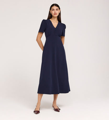 A woman with long brown hair stands against a plain background wearing the Nieve Navy Crepe Midi Dress, a V-neck, short-sleeved, empire waist midi with buttons on the bodice and pointed dark shoes.