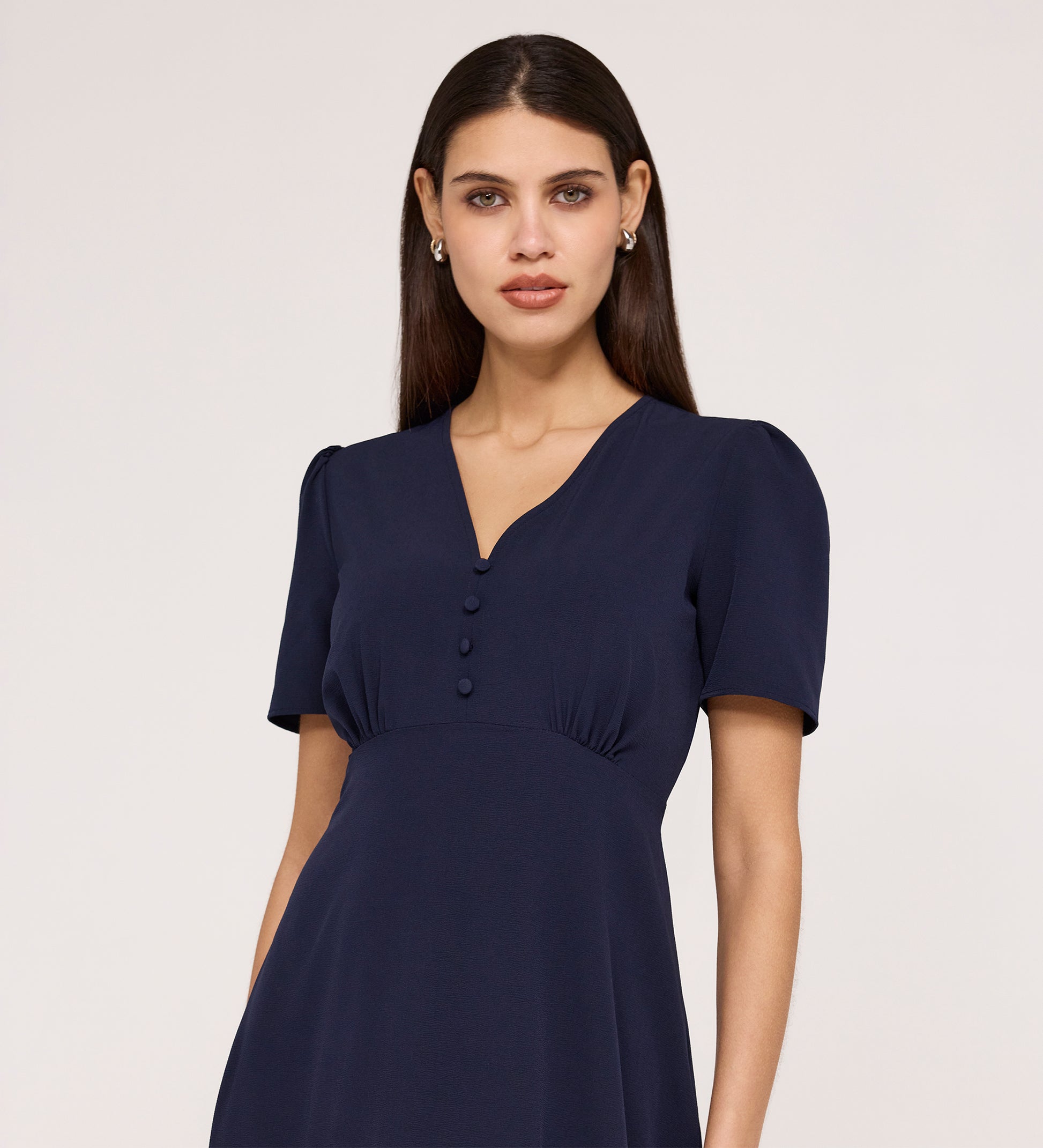 A woman with long brown hair wears the Nieve Navy Crepe Midi Dress, featuring an empire waist, V-neckline, and button details, while standing against a plain light background.