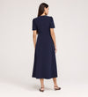 A woman with long brown hair stands facing away, wearing the Nieve Navy Crepe Midi Dress with a V-neckline and empire waist midi design, paired with open-toed heeled sandals against a plain light background.