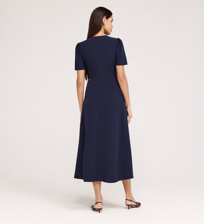 A woman with long brown hair stands facing away, wearing the Nieve Navy Crepe Midi Dress with a V-neckline and empire waist midi design, paired with open-toed heeled sandals against a plain light background.