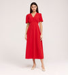 A woman with straight dark hair wears the Nieve Red Crepe Midi Dress, which has an empire waist, short sleeves, and a buttoned V-neck bodice. She stands against a plain background and pairs it with beige low-heeled shoes.
