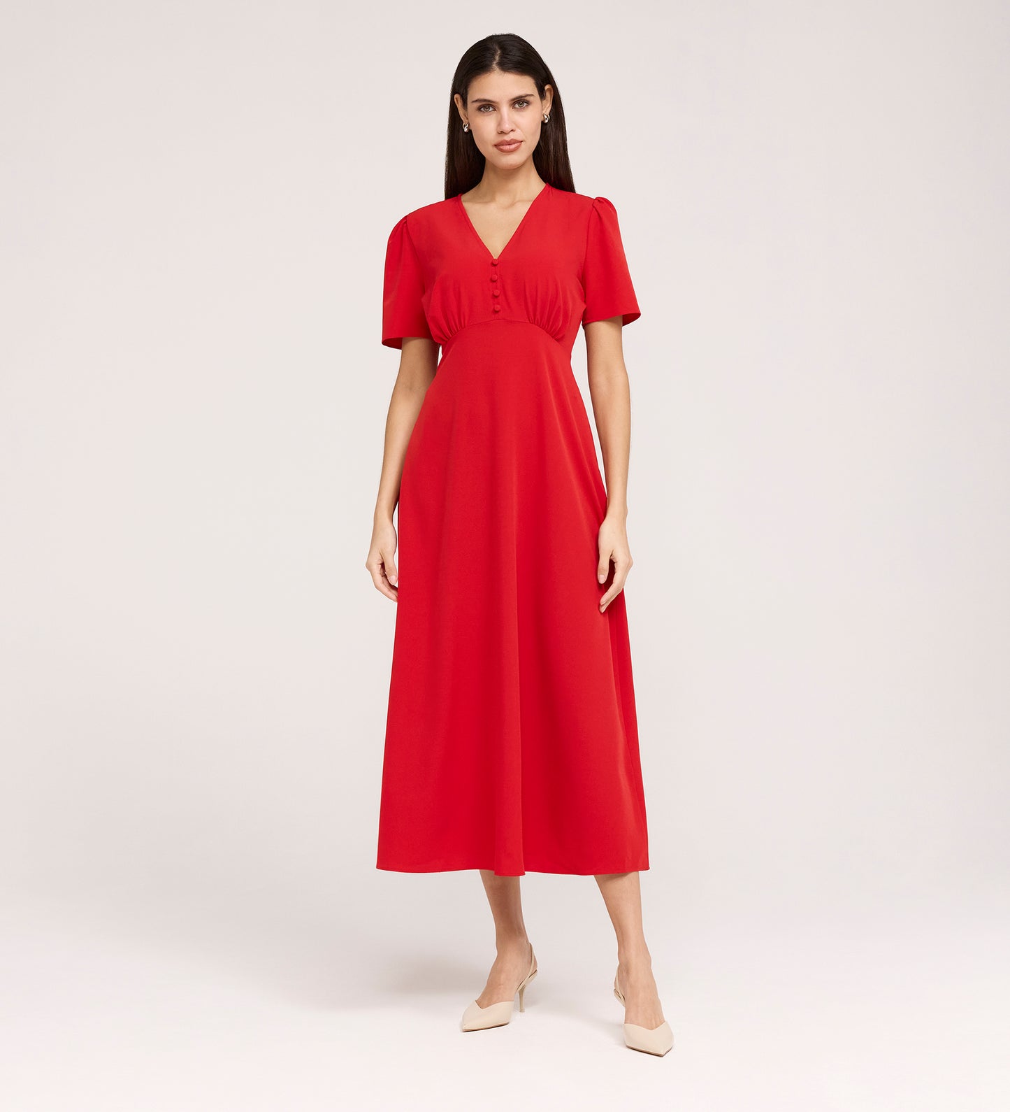 A woman with straight dark hair wears the Nieve Red Crepe Midi Dress, which has an empire waist, short sleeves, and a buttoned V-neck bodice. She stands against a plain background and pairs it with beige low-heeled shoes.