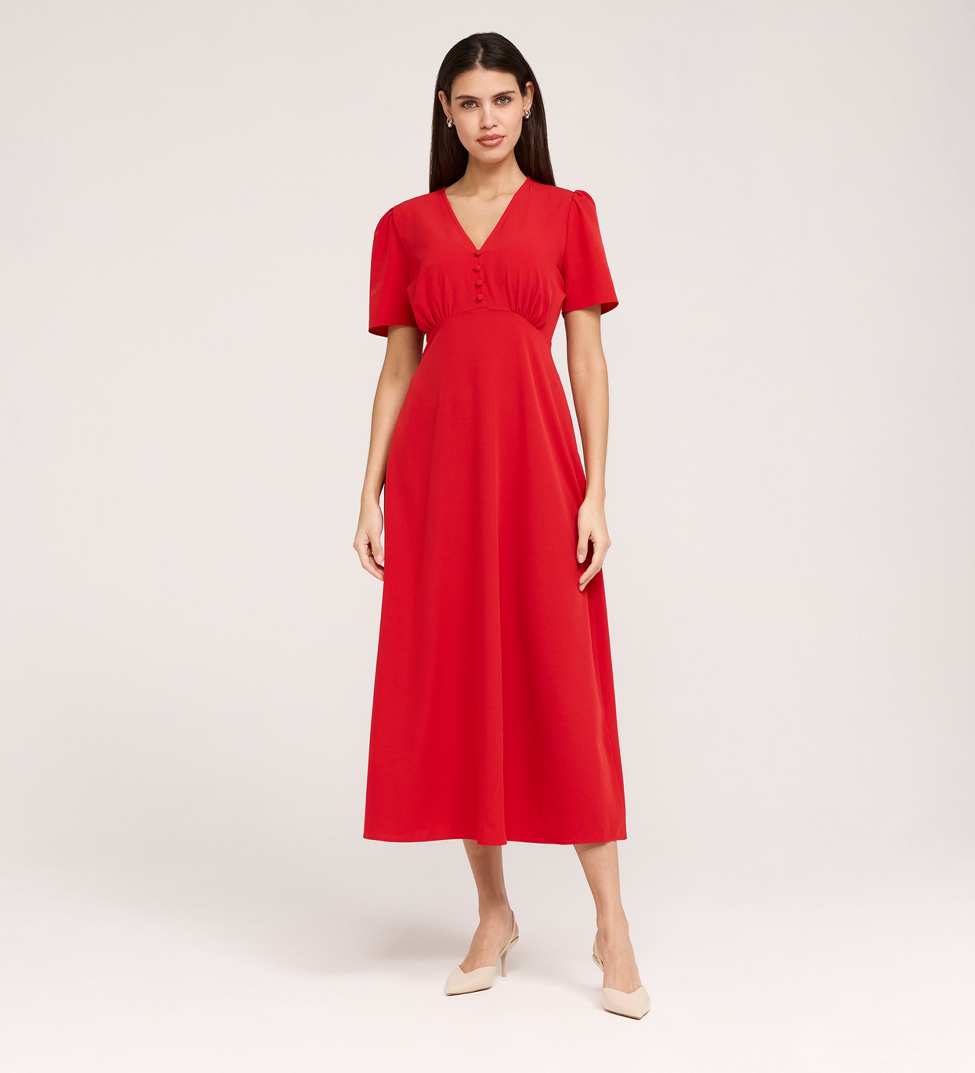 A woman with straight dark hair wears the Nieve Red Crepe Midi Dress, which has an empire waist, short sleeves, and a buttoned V-neck bodice. She stands against a plain background and pairs it with beige low-heeled shoes.