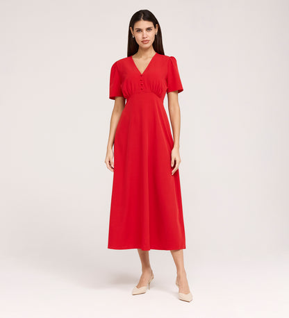 A woman with straight dark hair wears the Nieve Red Crepe Midi Dress, which has an empire waist, short sleeves, and a buttoned V-neck bodice. She stands against a plain background and pairs it with beige low-heeled shoes.