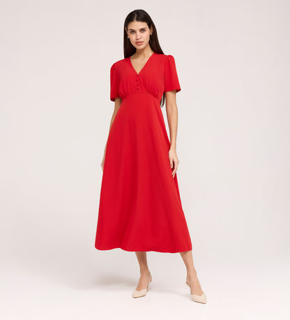 A woman stands against a plain background wearing the Nieve Red Crepe Midi Dress, a flowing midi-length dress with an empire waist, short sleeves, V-neck, front buttons, and beige pointed-toe low heels.
