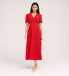 A woman stands against a plain background wearing the Nieve Red Crepe Midi Dress, a short-sleeve, bright red midi dress with an empire waist, buttoned bodice, and beige pointed-toe shoes.