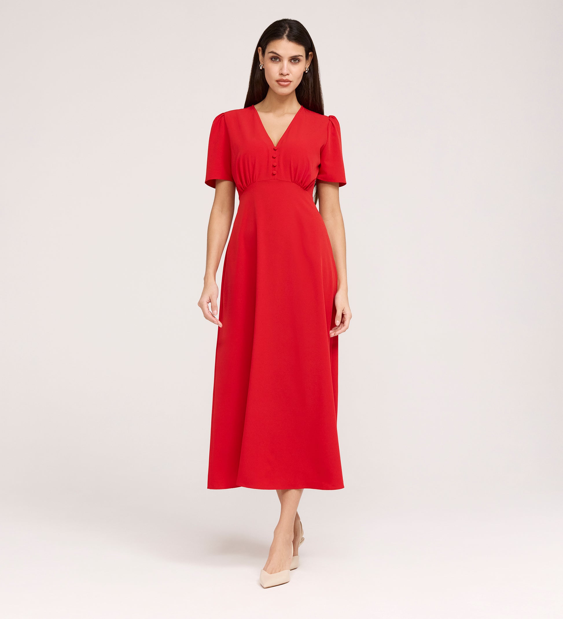 A woman stands against a plain background wearing the Nieve Red Crepe Midi Dress, a short-sleeve, bright red midi dress with an empire waist, buttoned bodice, and beige pointed-toe shoes.
