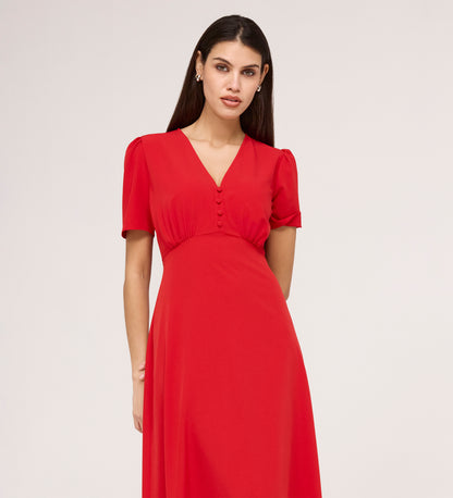 A woman with long dark hair stands against a plain light background, wearing the Nieve Red Crepe Midi Dress—an empire waist, short-sleeved V-neck dress with buttons on the bodice.