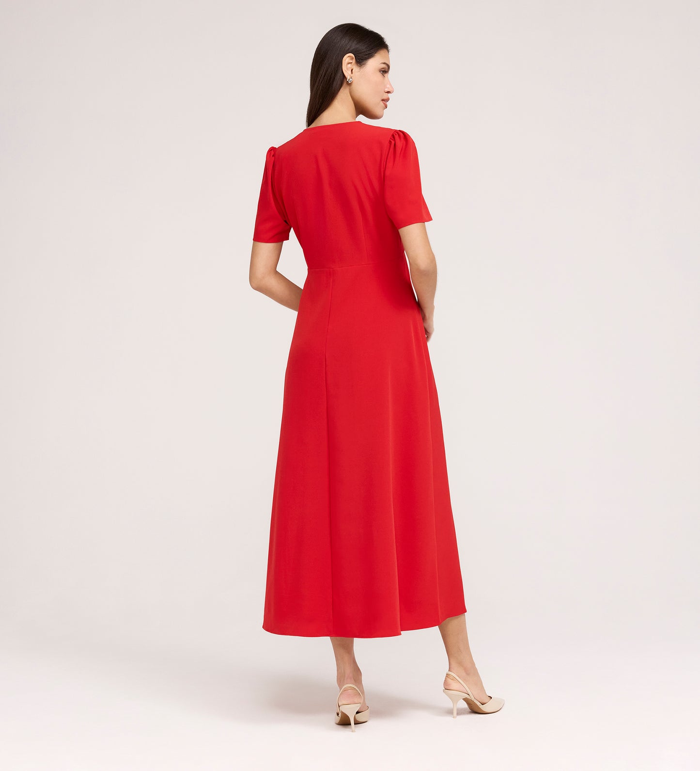 A woman faces away, wearing the Nieve Red Crepe Midi Dress with a square neckline and empire waist. The short-sleeved, midi dress pairs elegantly with beige heeled sandals against a plain light background.