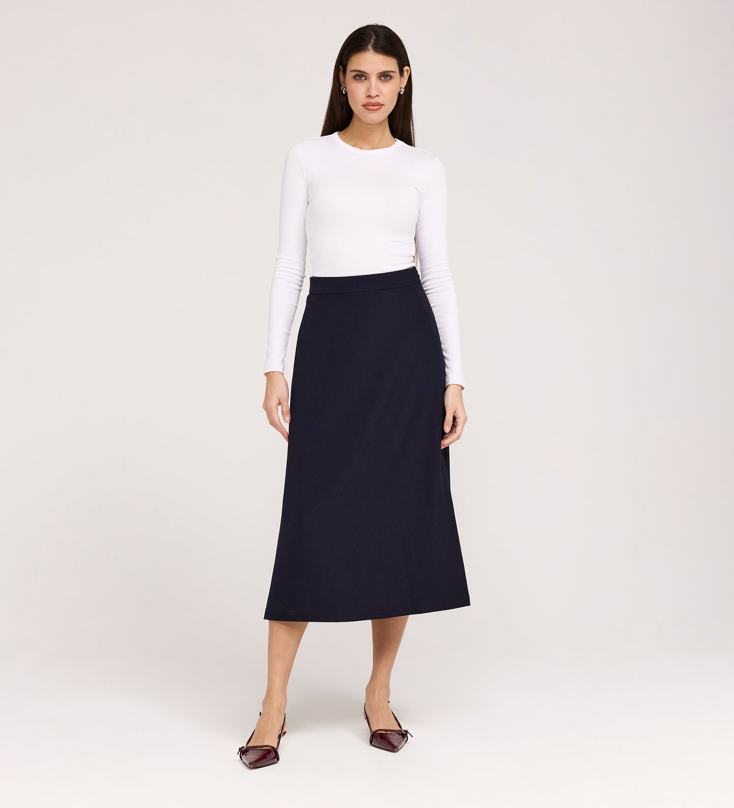 A woman stands against a plain background, wearing a fitted white long-sleeve top and the Ramona Navy Rich Crepe Midi Skirt, paired with pointed maroon flats. She faces forward with a neutral expression.