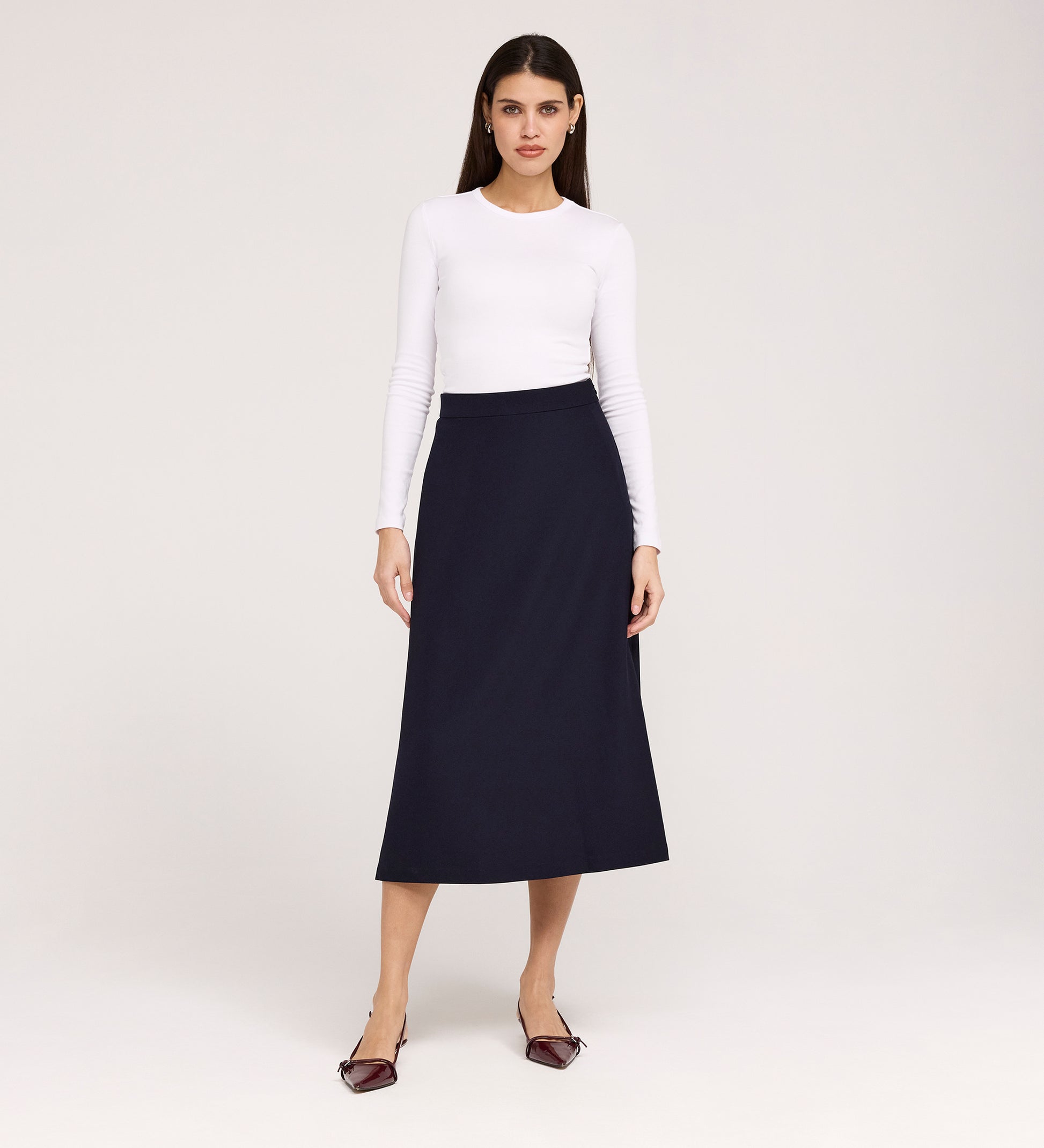 A woman stands against a plain background, wearing a fitted white long-sleeve top and the Ramona Navy Rich Crepe Midi Skirt, paired with pointed maroon flats. She faces forward with a neutral expression.