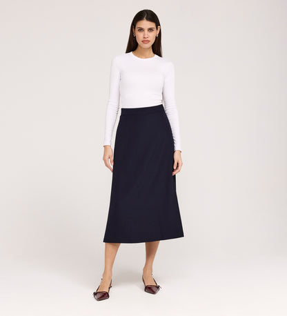 A woman stands against a plain background, wearing a fitted white long-sleeve top and the Ramona Navy Rich Crepe Midi Skirt, paired with pointed maroon flats. She faces forward with a neutral expression.