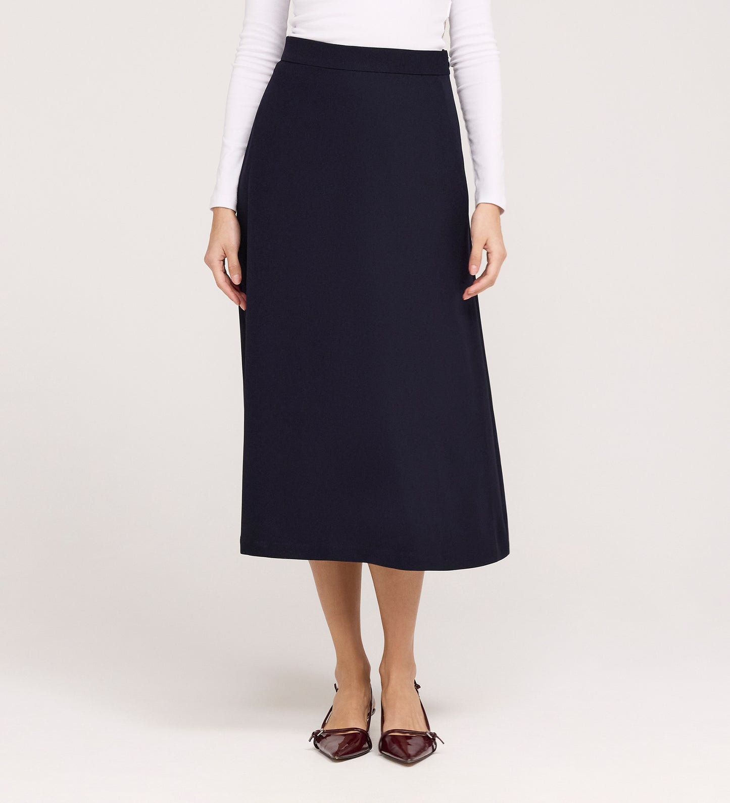A woman, her face not visible, stands against a light background wearing the Ramona Navy Rich Crepe Midi Skirt paired with a fitted white long-sleeve top and pointed maroon flats.