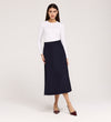 A woman stands against a plain background wearing a long-sleeved white top, pointed burgundy shoes, and the Ramona Navy Rich Crepe Midi Skirt. She faces forward with a neutral expression and relaxed posture.