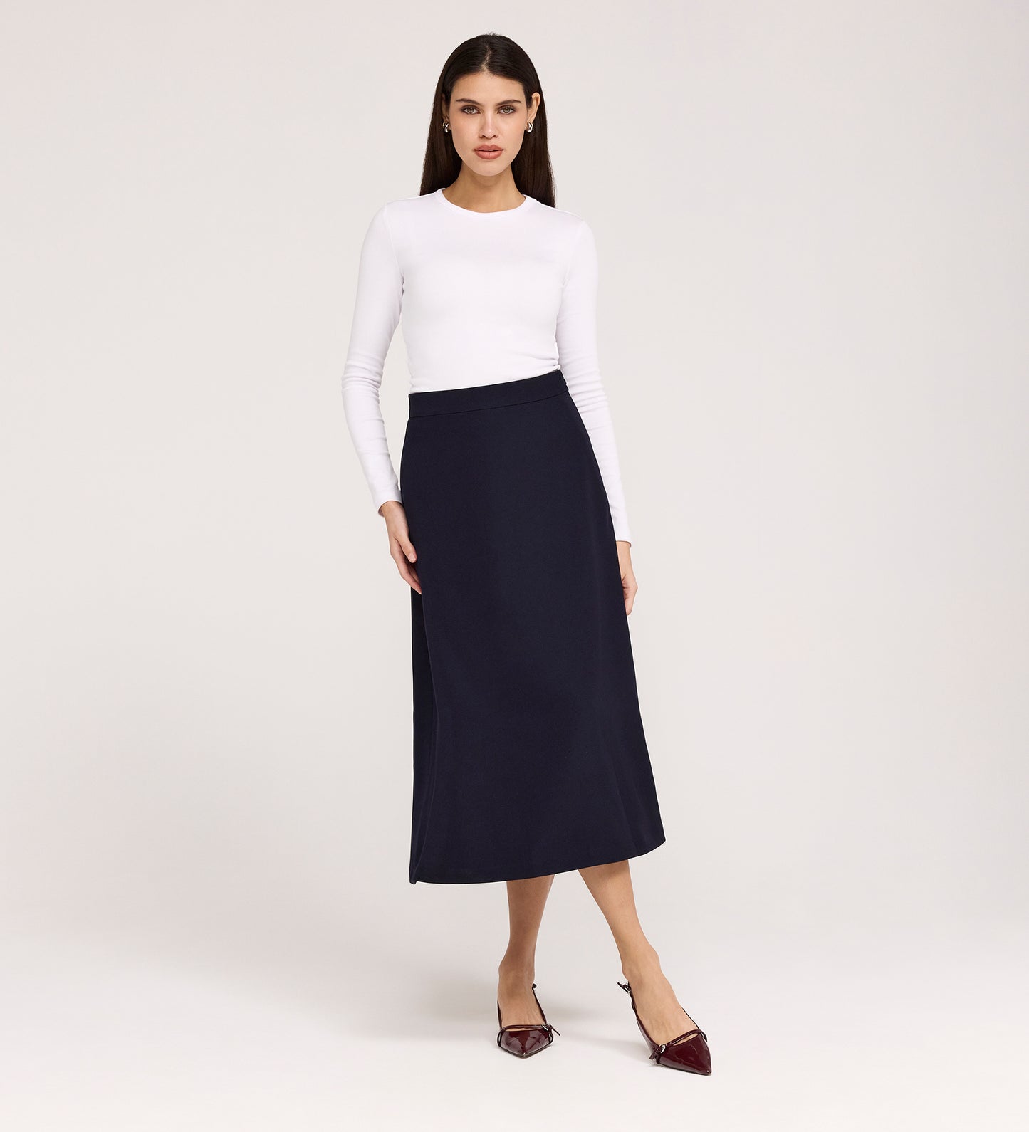 A woman stands against a plain background wearing a long-sleeved white top, pointed burgundy shoes, and the Ramona Navy Rich Crepe Midi Skirt. She faces forward with a neutral expression and relaxed posture.