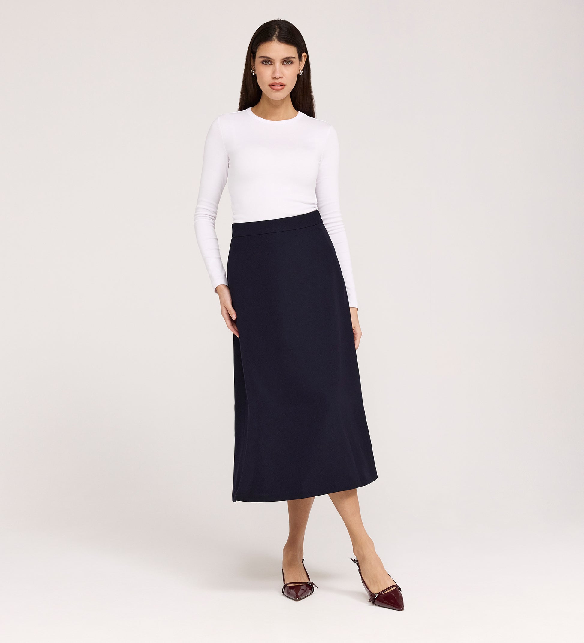 A woman stands against a plain background wearing a long-sleeved white top, pointed burgundy shoes, and the Ramona Navy Rich Crepe Midi Skirt. She faces forward with a neutral expression and relaxed posture.
