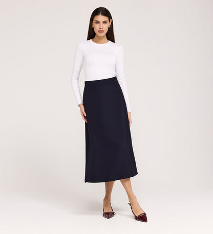 A woman stands against a plain background wearing a long-sleeved white top, pointed burgundy shoes, and the Ramona Navy Rich Crepe Midi Skirt. She faces forward with a neutral expression and relaxed posture.