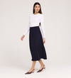 A woman stands against a plain light background, looking to the side, wearing a white long-sleeve top, the Ramona Navy Rich Crepe Midi Skirt, and burgundy low-heeled shoes.