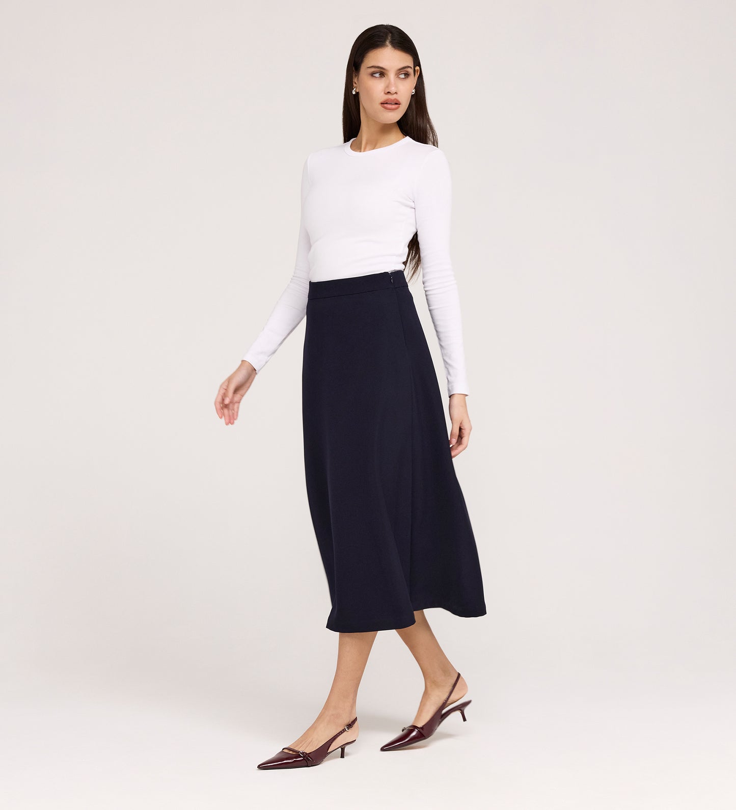 A woman stands against a plain light background, looking to the side, wearing a white long-sleeve top, the Ramona Navy Rich Crepe Midi Skirt, and burgundy low-heeled shoes.