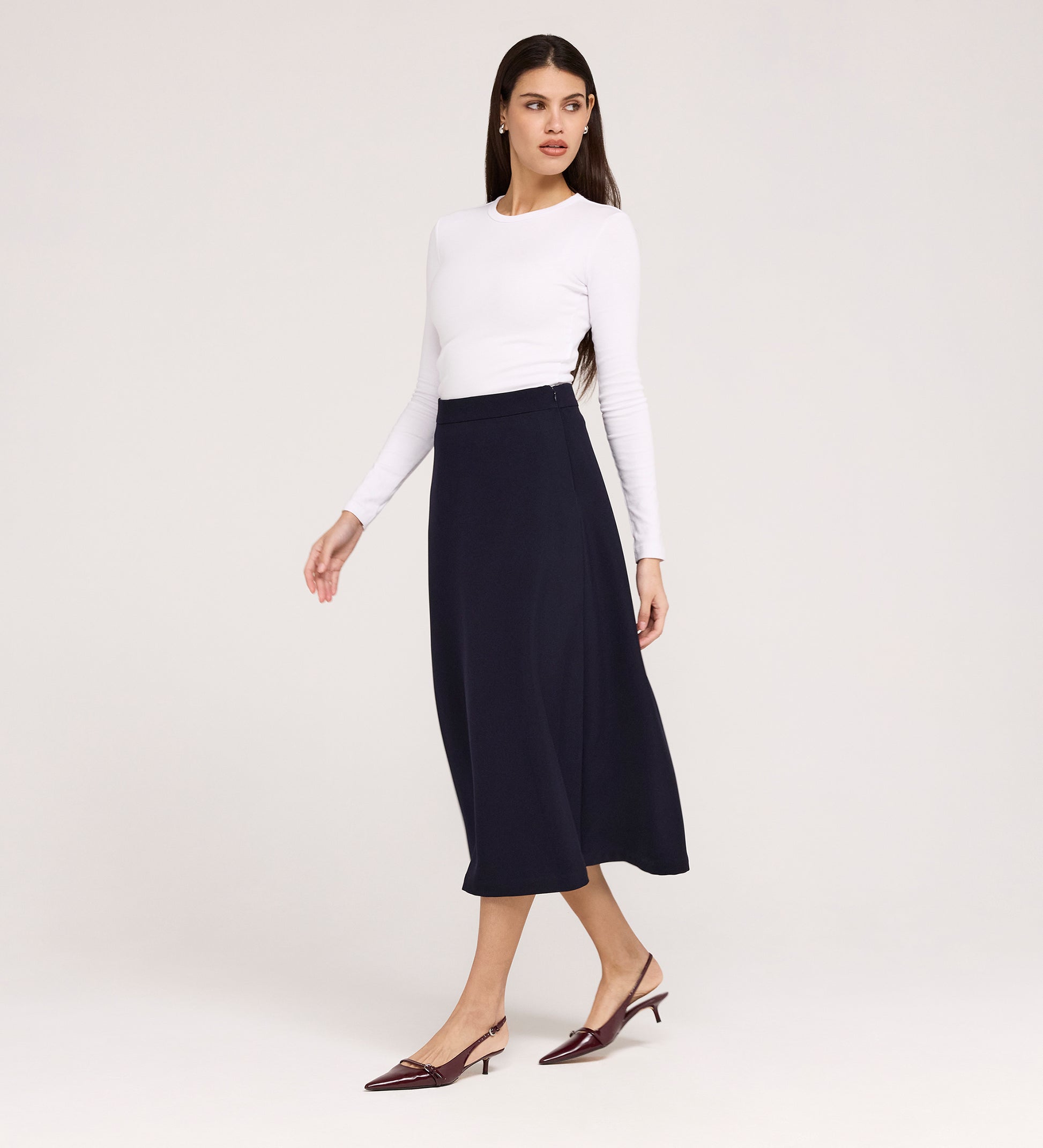 A woman stands against a plain light background, looking to the side, wearing a white long-sleeve top, the Ramona Navy Rich Crepe Midi Skirt, and burgundy low-heeled shoes.