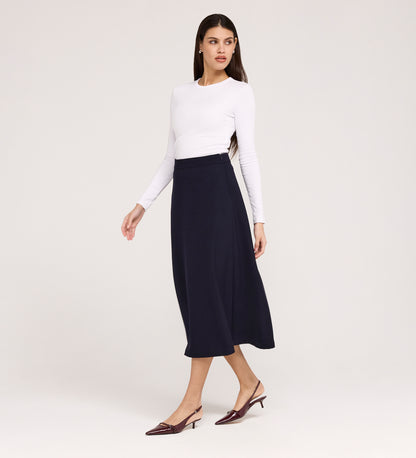 A woman stands against a plain light background, looking to the side, wearing a white long-sleeve top, the Ramona Navy Rich Crepe Midi Skirt, and burgundy low-heeled shoes.