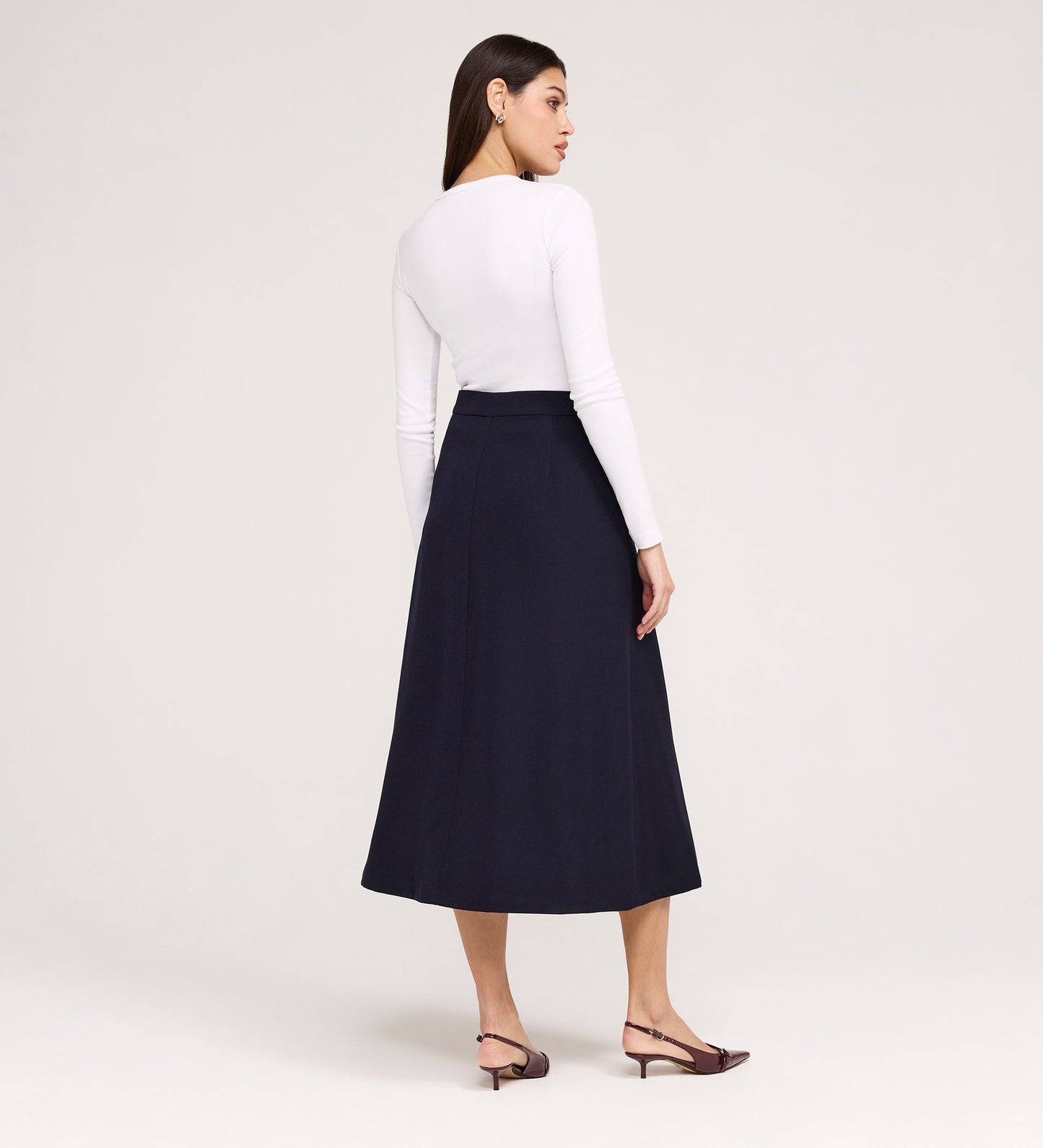 A woman stands with her back to the camera, wearing a fitted long-sleeve white top, the Ramona Navy Rich Crepe Midi Skirt, and brown low-heeled slingbacks against a plain light background.