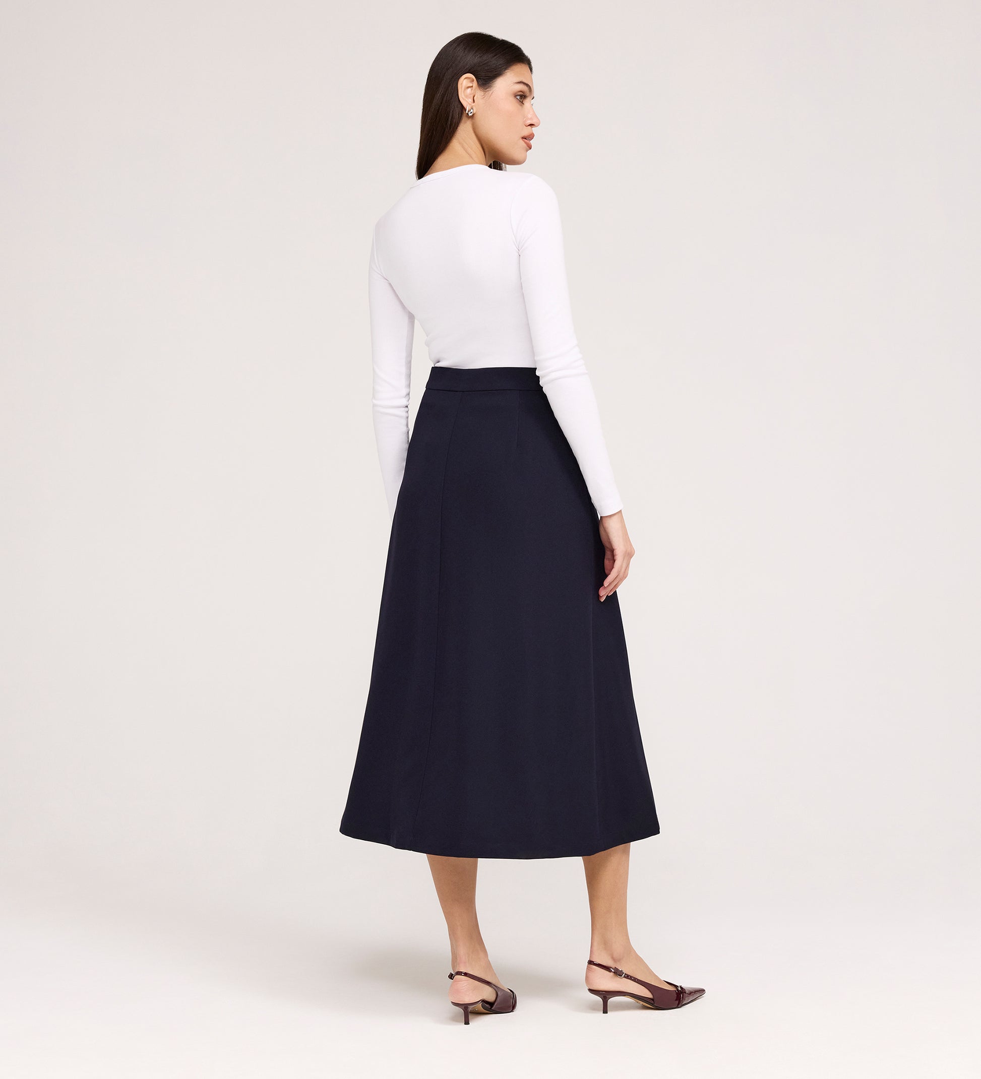 A woman stands with her back to the camera, wearing a fitted long-sleeve white top, the Ramona Navy Rich Crepe Midi Skirt, and brown low-heeled slingbacks against a plain light background.
