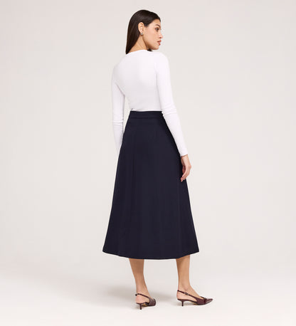 A woman stands with her back to the camera, wearing a fitted long-sleeve white top, the Ramona Navy Rich Crepe Midi Skirt, and brown low-heeled slingbacks against a plain light background.
