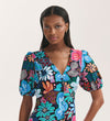 A woman with long, straight black hair wears the Sarah Blue Floral Crepe Midi Dress with a vibrant abstract floral pattern, v-neckline, short puff sleeves, and button-through front. She stands against a plain background, looking at the camera neutrally.