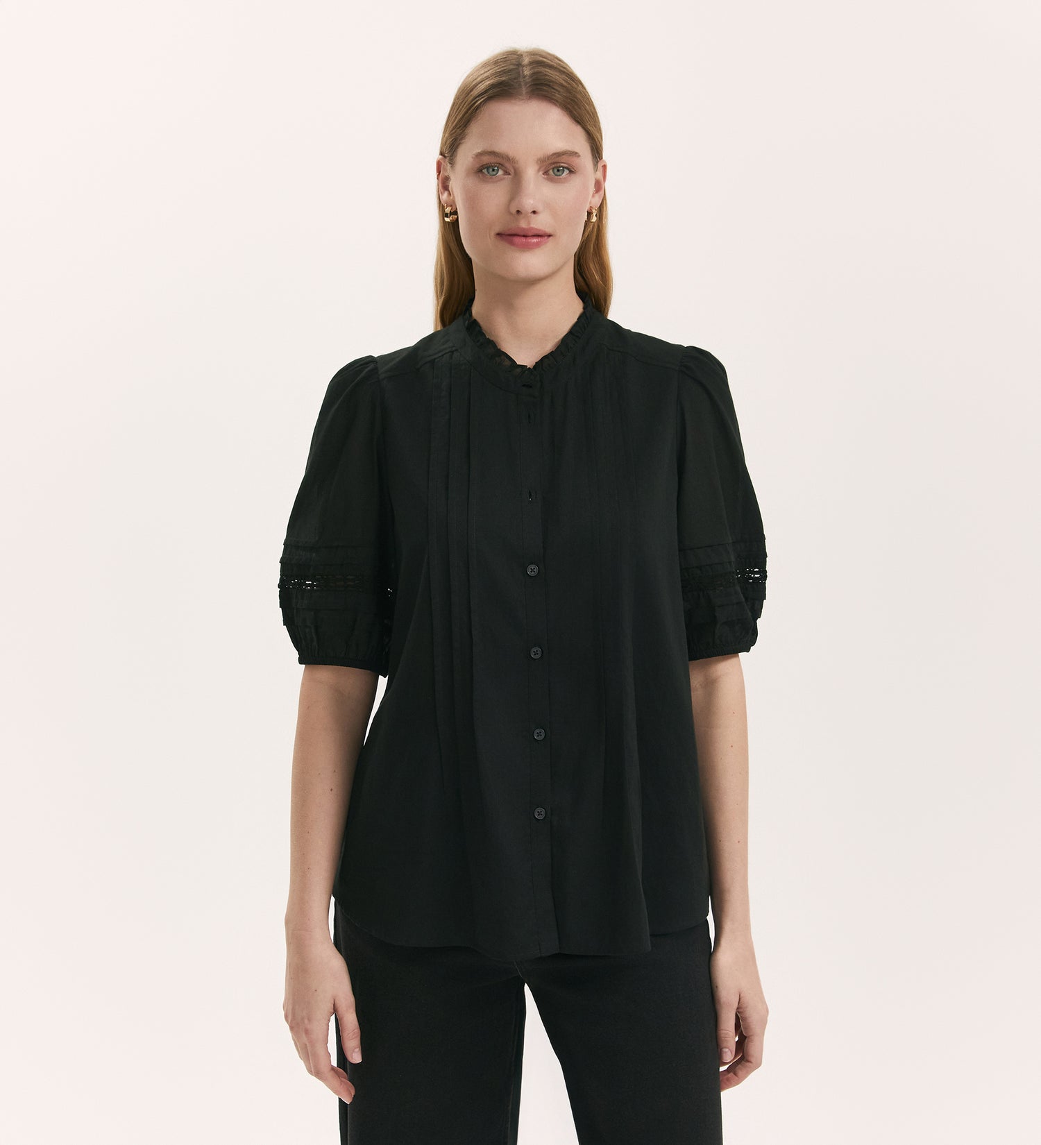 A woman with straight, light brown hair models the Adalia Black Cotton Blouse, a short-sleeved top with puffed sleeves and pintuck details. She stands facing forward against a plain, light background.