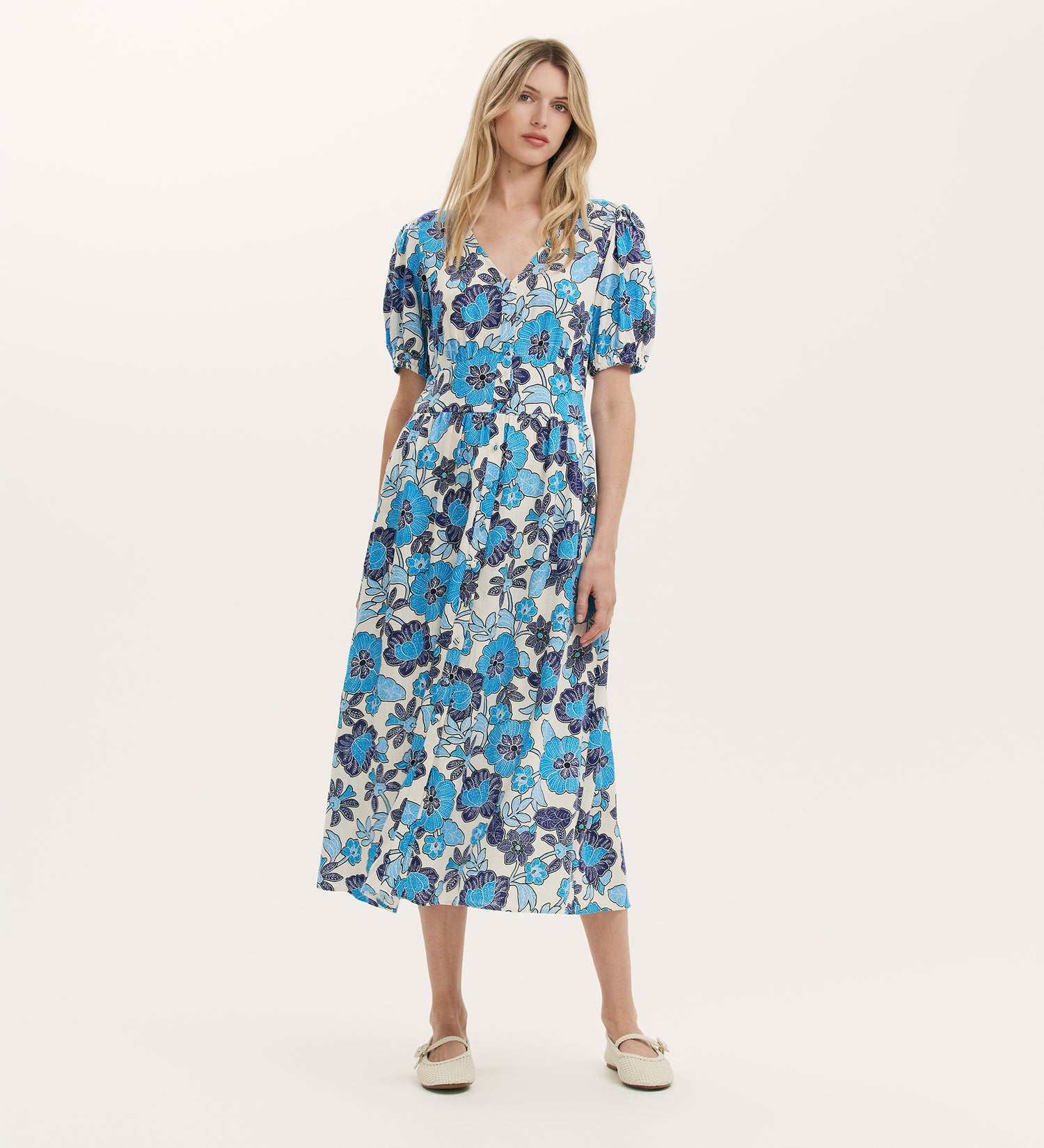 A woman wears the Addison Blue Flower Midi Dress with Linen, featuring a blue and white floral print, puff sleeves, V-neck, belted waist, and pockets, paired with white flats against a plain light background.