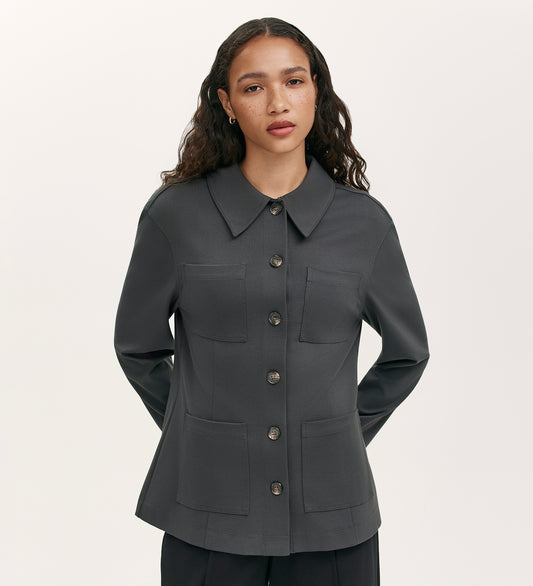 A woman with long curly hair models the Agness Charcoal Ponte Jersey Button Down Jacket, featuring a relaxed fit, pointed collar, and four front pockets, against a plain light background with her hands behind her back.