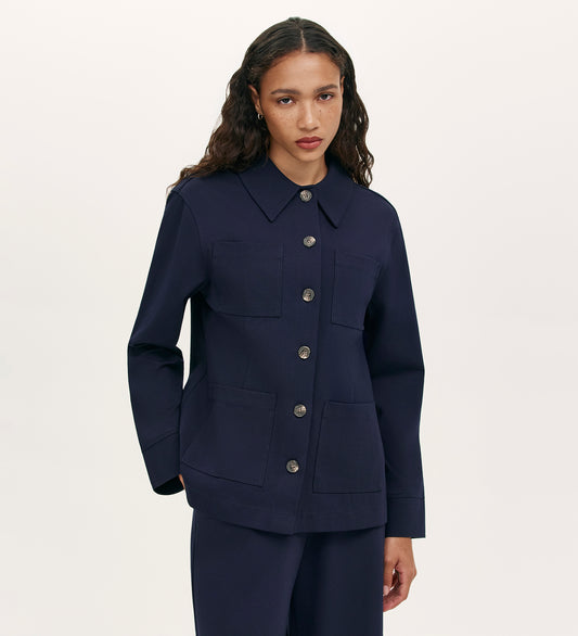 A woman with wavy brown hair wears the Agness Navy Ponte Jersey Button Down Jacket, a relaxed-fit style with four large front pockets and matching pants, set against a plain light background.