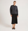 A woman with her hair tied back stands against a plain background wearing the Ailsa Charcoal Knitted Cardigan over a black midi skirt and flat shoes, looking straight ahead with a neutral expression—showcasing this versatile layering piece.