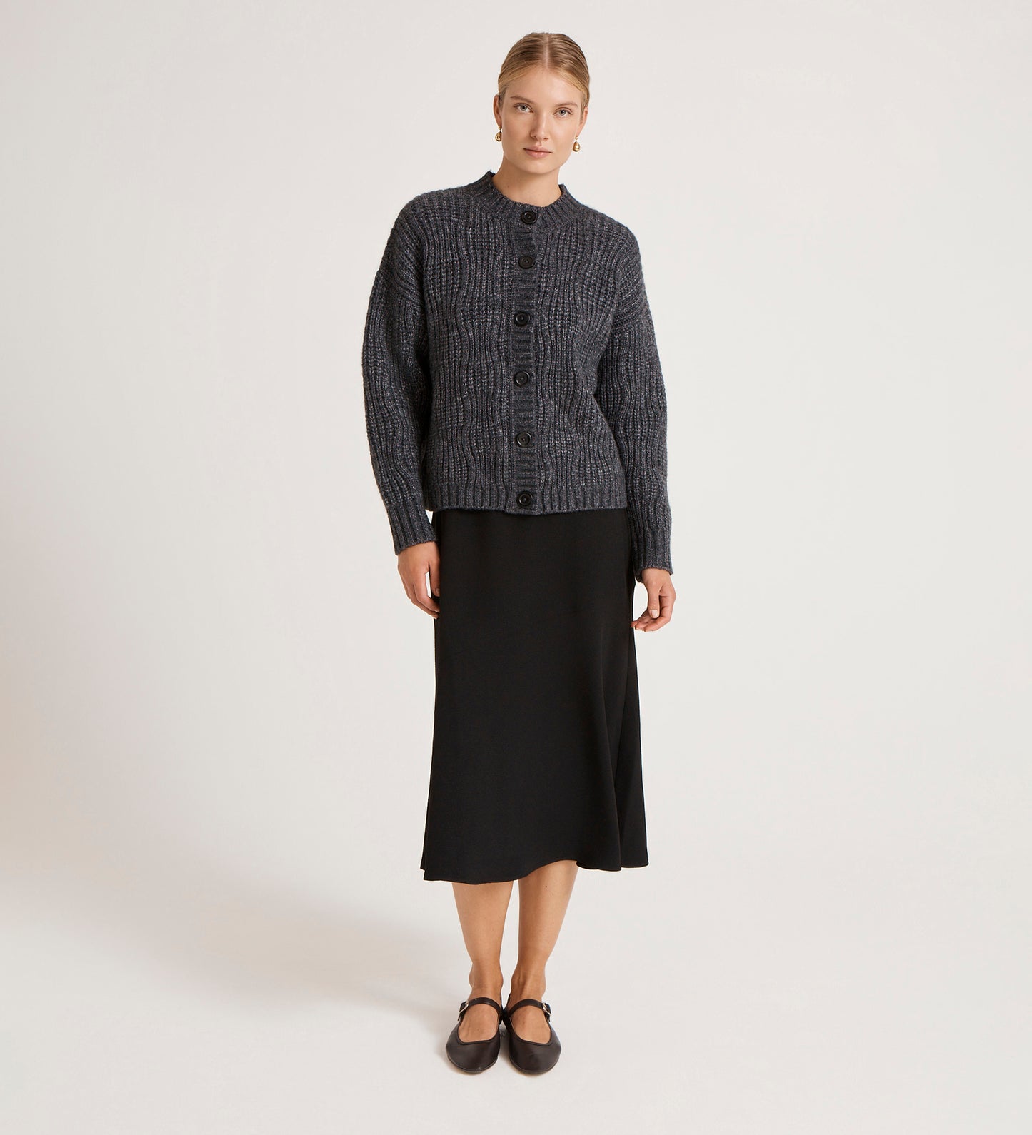 A woman with her hair tied back stands against a plain background wearing the Ailsa Charcoal Knitted Cardigan over a black midi skirt and flat shoes, looking straight ahead with a neutral expression—showcasing this versatile layering piece.