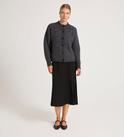 A woman with her hair tied back stands against a plain background wearing the Ailsa Charcoal Knitted Cardigan over a black midi skirt and flat shoes, looking straight ahead with a neutral expression—showcasing this versatile layering piece.