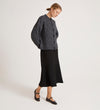 A woman stands against a plain light background wearing the Ailsa Charcoal Knitted Cardigan—an effortless essential—paired with a black midi skirt and Mary Jane shoes, her hair pulled back as she looks downward with her hands behind her back.