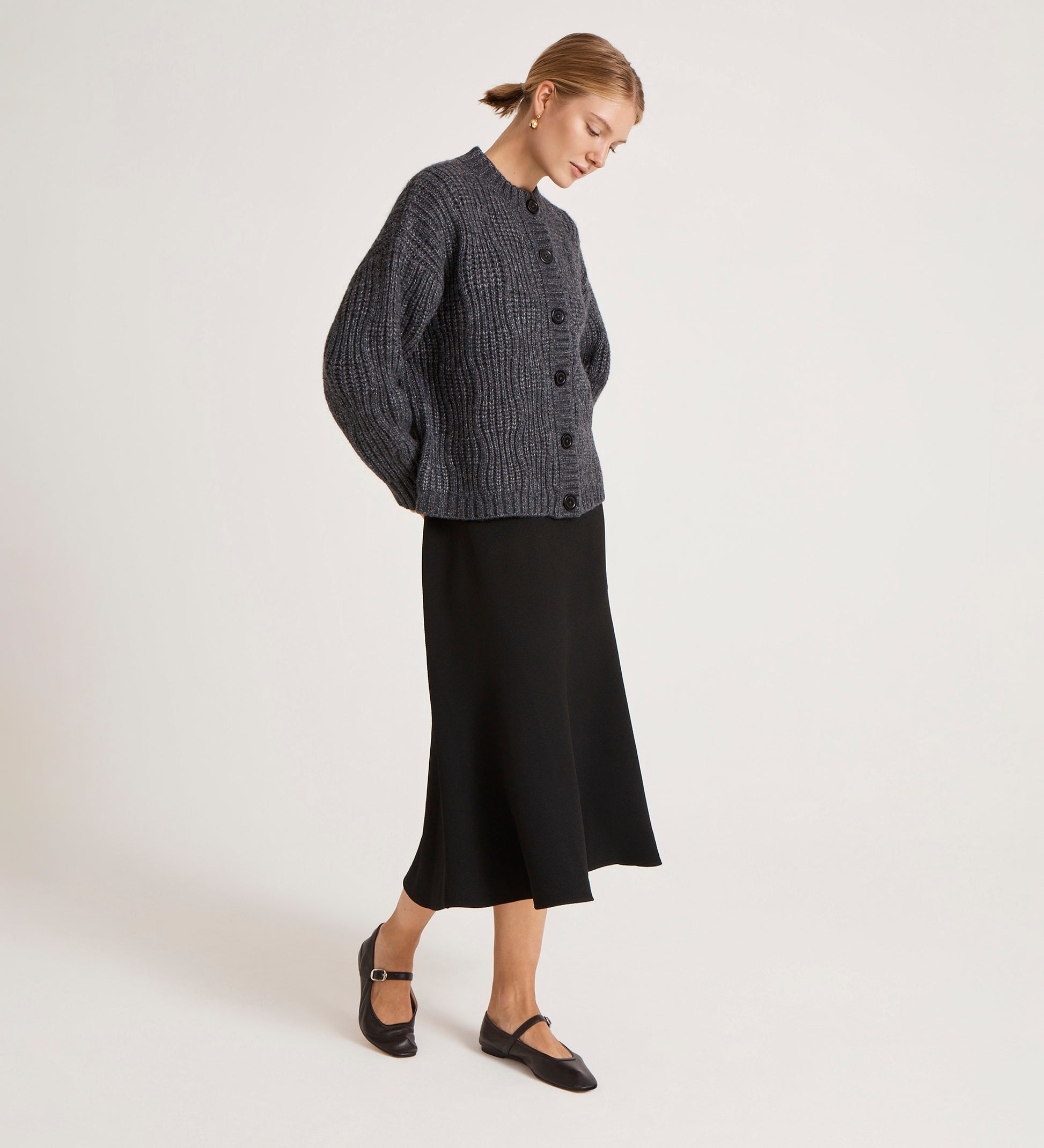A woman stands against a plain light background wearing the Ailsa Charcoal Knitted Cardigan—an effortless essential—paired with a black midi skirt and Mary Jane shoes, her hair pulled back as she looks downward with her hands behind her back.