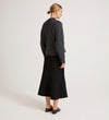 A woman with a low blonde bun faces away, showcasing everyday layering in the Ailsa Charcoal Knitted Cardigan paired with a black midi skirt and black shoes, set against a light background.