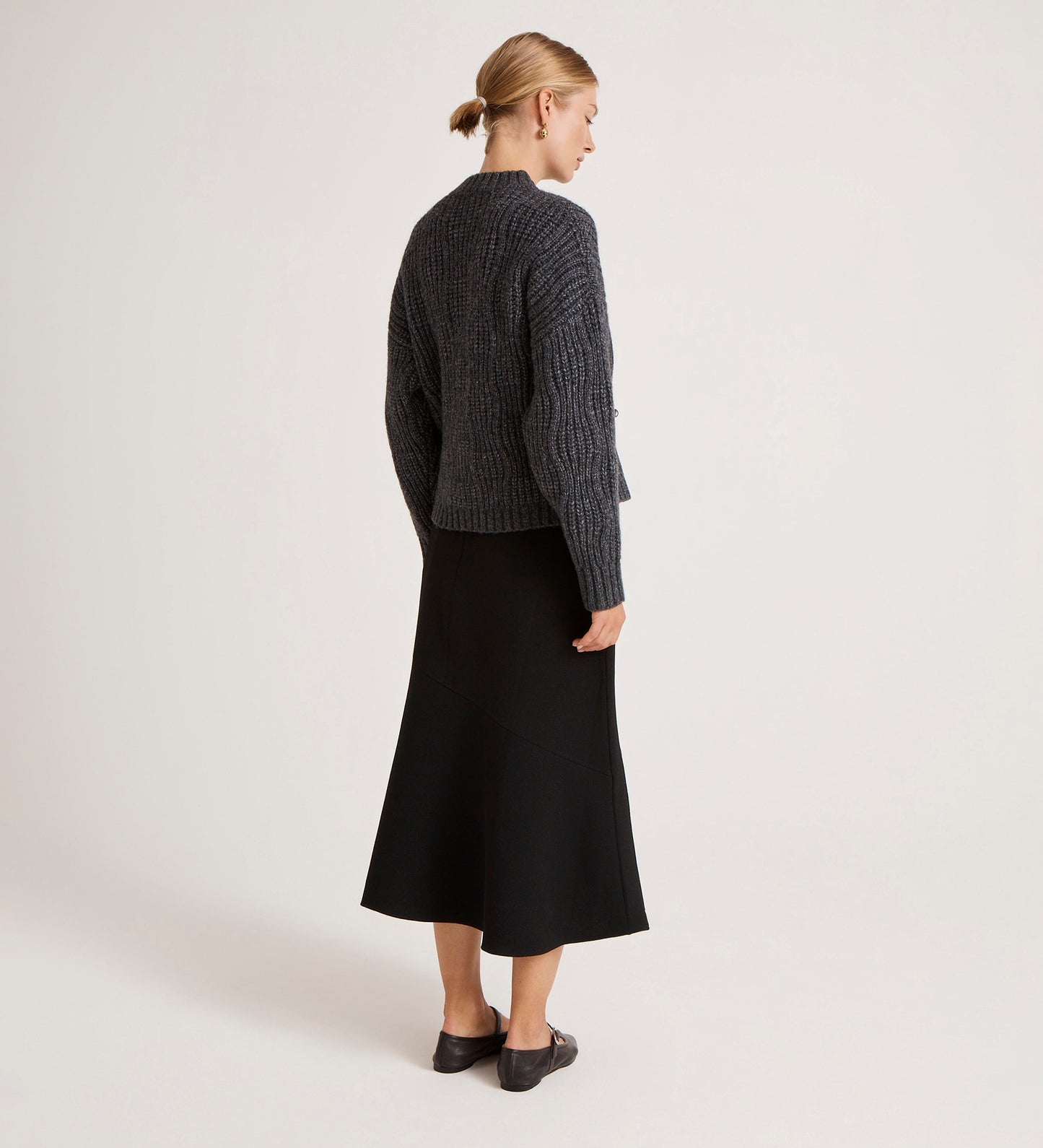 A woman with a low blonde bun faces away, showcasing everyday layering in the Ailsa Charcoal Knitted Cardigan paired with a black midi skirt and black shoes, set against a light background.
